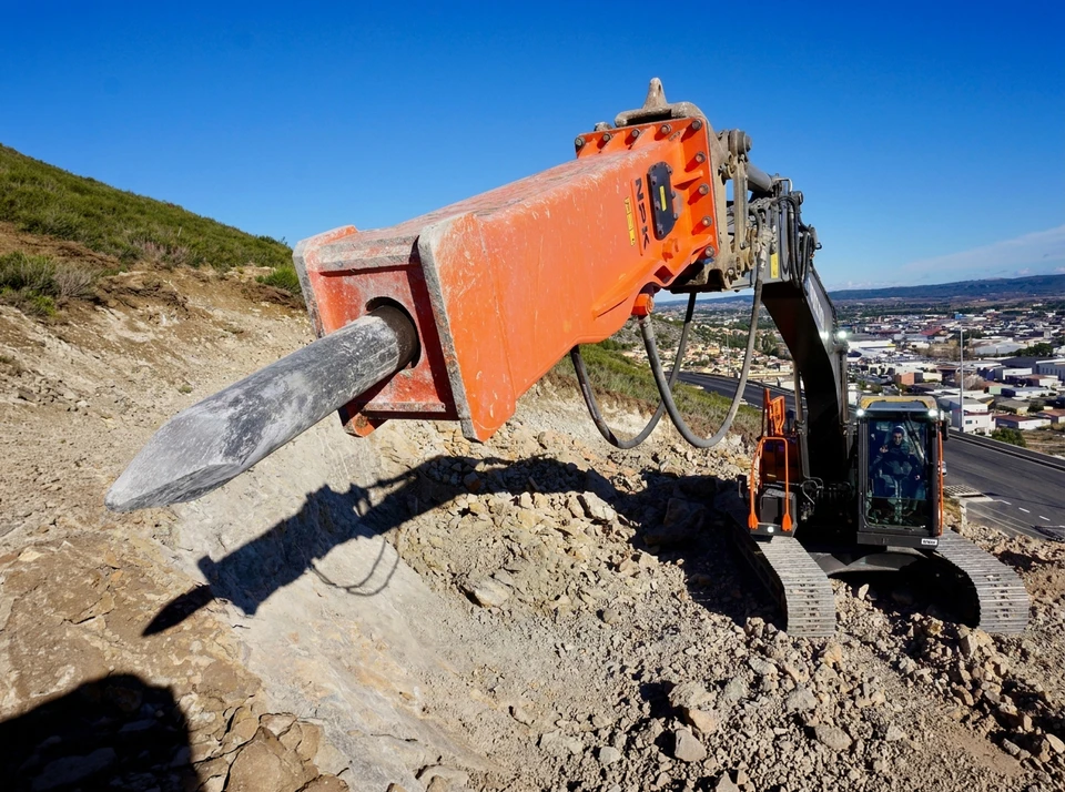 Pelleteuse hydraulique jaune sur chenilles en action sur chantier extérieur de terrassement, opérateur visible dans la cabine vitrée relevant le godet rempli de terre, sol argileux excavé en arrière-plan, ciel dégagé, image de qualité professionnelle
